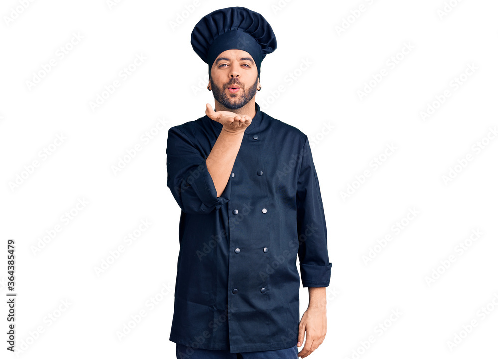 Young hispanic man wearing cooker uniform looking at the camera blowing a kiss with hand on air being lovely and sexy. love expression. Young hispanic man wearing cooker uniform looking at the camera blowing a kiss with hand on air being lovely and sexy. love expression.