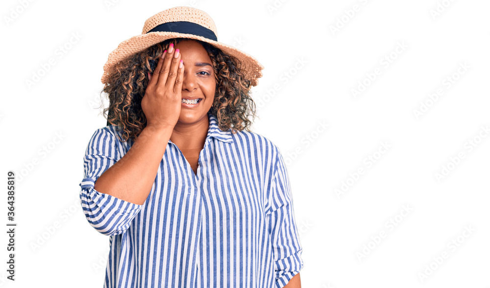 Young african american plus size woman wearing summer hat covering one eye with hand, confident smile on face and surprise emotion. Young african american plus size woman wearing summer hat covering one eye with hand, confident smile on face and surprise emotion.