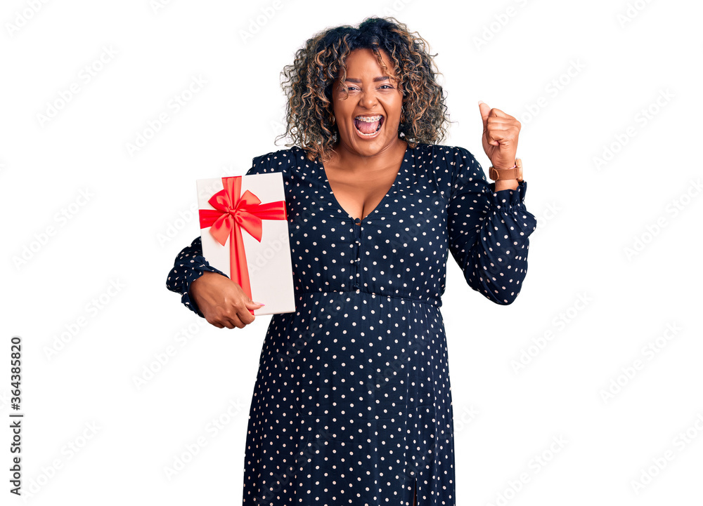 Young african american plus size woman holding gift screaming proud, celebrating victory and success very excited with raised arms Young african american plus size woman holding gift screaming proud, celebrating victory and success very excited with raised arms