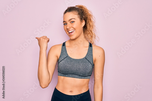 Young beautiful blonde sportswoman with blue eyes doing exercise wearing sportswear smiling with happy face looking and pointing to the side with thumb up. © Krakenimages.com