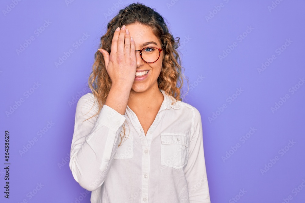 Young beautiful woman with blue eyes wearing casual shirt and glasses over purple background covering one eye with hand, confident smile on face and surprise emotion.