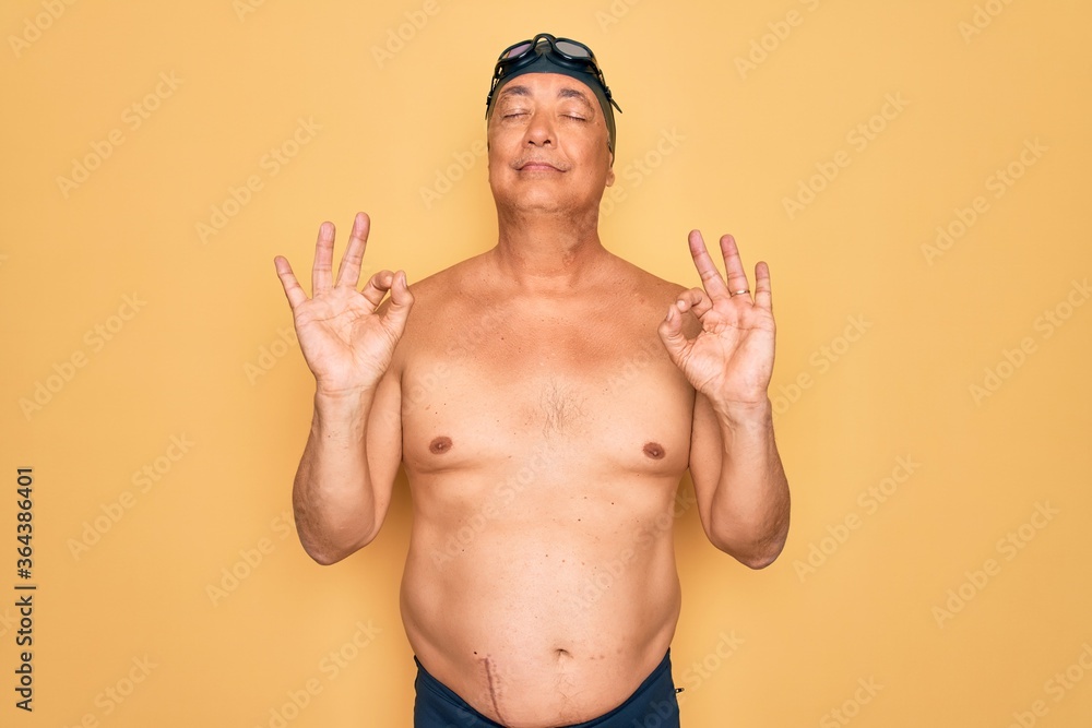 Middle age senior grey-haired swimmer man wearing swimsuit, cap and goggles relaxed and smiling with eyes closed doing meditation gesture with fingers. Yoga concept.