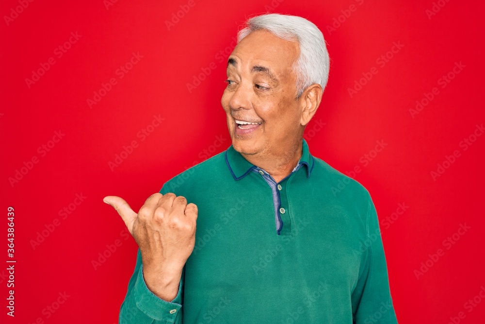 Fototapeta premium Middle age senior grey-haired man wearing casual sweater over red isoalted background smiling with happy face looking and pointing to the side with thumb up.