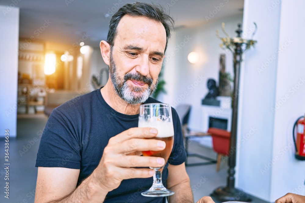 Middle age handsome man wearing casual tshirt smiling happy. Sitting with smile on face holding glass of beer at restaurant