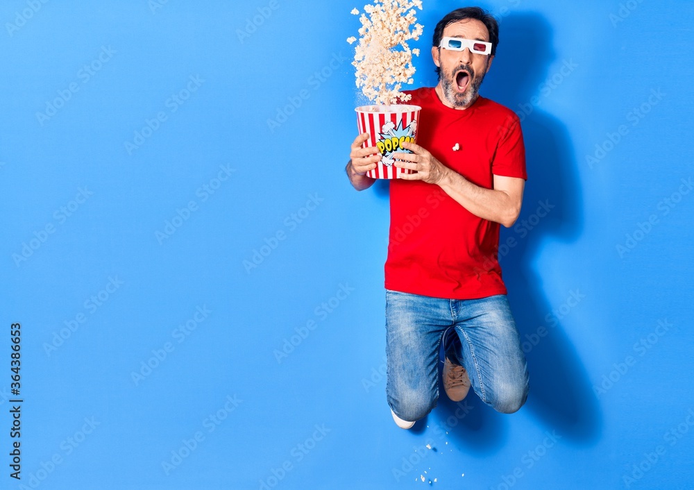 Middle age handsome hispanic man watching movie using 3d glasses. Holding bucket of popcorn jumping over isolated blue background Middle age handsome hispanic man watching movie using 3d glasses. Holding bucket of popcorn jumping over isolated blue background