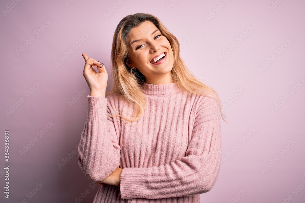Young beautiful blonde woman wearing casual pink sweater over isolated background with a big smile on face, pointing with hand and finger to the side looking at the camera. Young beautiful blonde woman wearing casual pink sweater over isolated background with a big smile on face, pointing with hand and finger to the side looking at the camera.