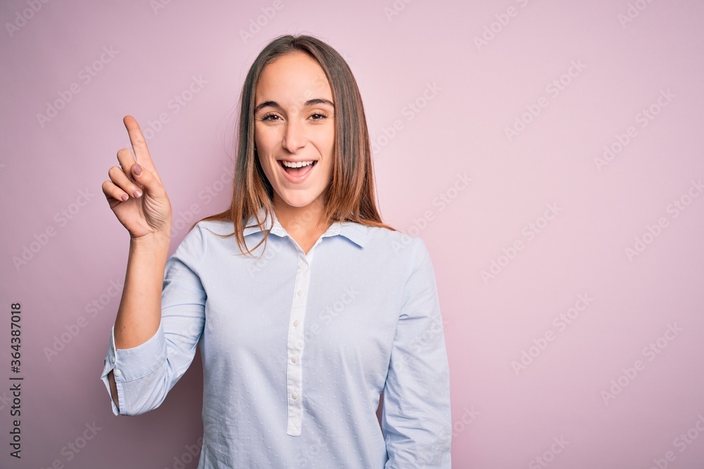 Young beautiful businesswoman wearing elegant shirt standing over isolated pink background showing and pointing up with finger number one while smiling confident and happy. Young beautiful businesswoman wearing elegant shirt standing over isolated pink background showing and pointing up with finger number one while smiling confident and happy.