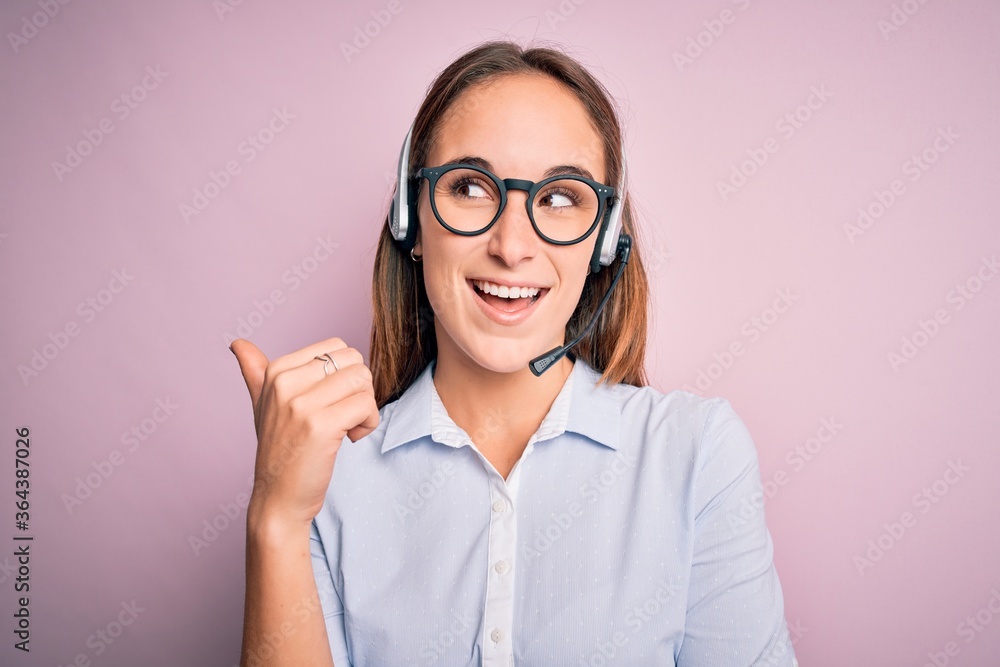 Young beautiful call center agent woman wearing glasses working using headset smiling with happy face looking and pointing to the side with thumb up. Young beautiful call center agent woman wearing glasses working using headset smiling with happy face looking and pointing to the side with thumb up.