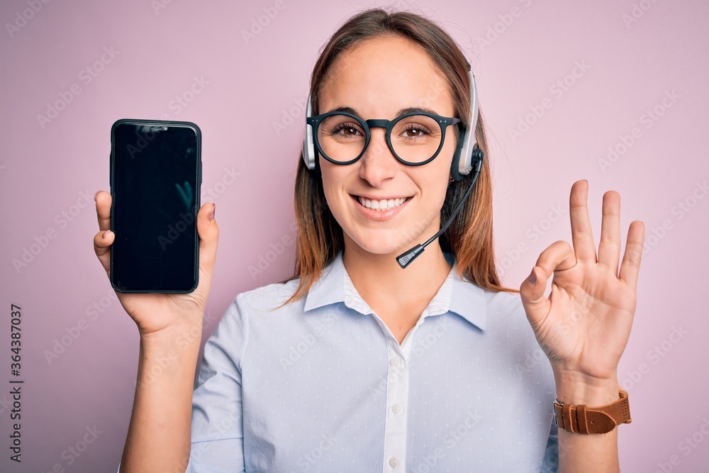 Beautiful call center agent woman working using headset holding smartphone holding screen doing ok sign with fingers, excellent symbol