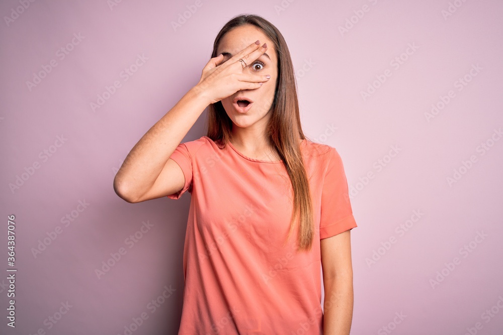 Young beautiful woman wearing casual t-shirt standing over isolated pink background peeking in shock covering face and eyes with hand, looking through fingers with embarrassed expression. Young beautiful woman wearing casual t-shirt standing over isolated pink background peeking in shock covering face and eyes with hand, looking through fingers with embarrassed expression.