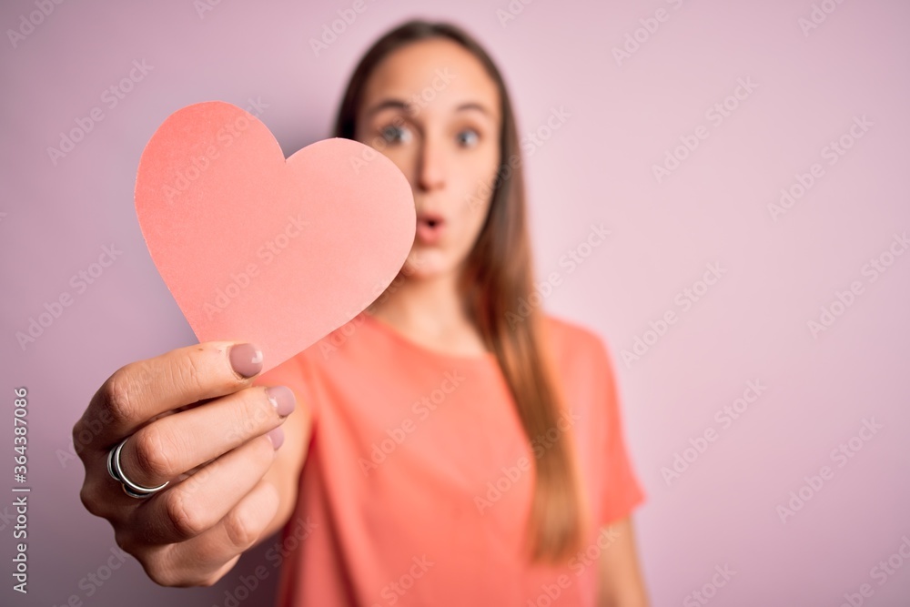 Young beautiful romantic woman holding paper heart shape over isolated pink background scared in shock with a surprise face, afraid and excited with fear expression Young beautiful romantic woman holding paper heart shape over isolated pink background scared in shock with a surprise face, afraid and excited with fear expression