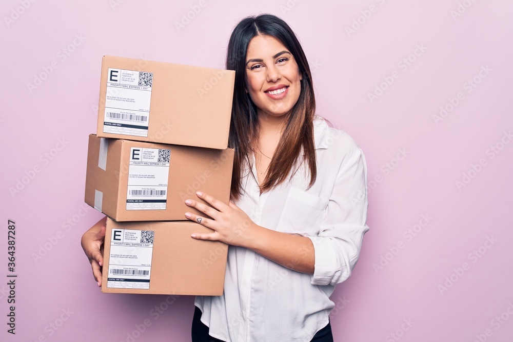 Young beautiful brunette woman holding deliver packages standing over pink background looking positive and happy standing and smiling with a confident smile showing teeth Young beautiful brunette woman holding deliver packages standing over pink background looking positive and happy standing and smiling with a confident smile showing teeth