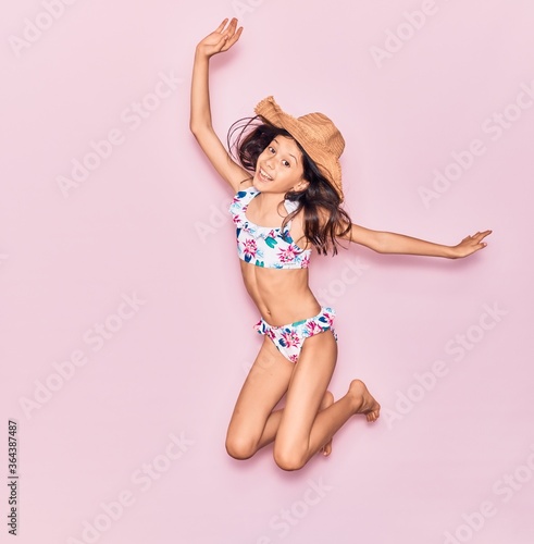 Adorable hispanic child girl on vacation wearing bikini and hat smiling happy. Jumping with smile on face over isolated pink background