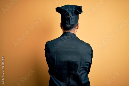 Young brazilian chef man wearing cooker uniform and hat over isolated yellow background standing backwards looking away with crossed arms