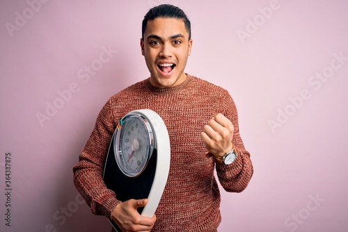 Young brazilian man doing diet to lose weigth holding scale over isolated pink background screaming proud and celebrating victory and success very excited, cheering emotion photo