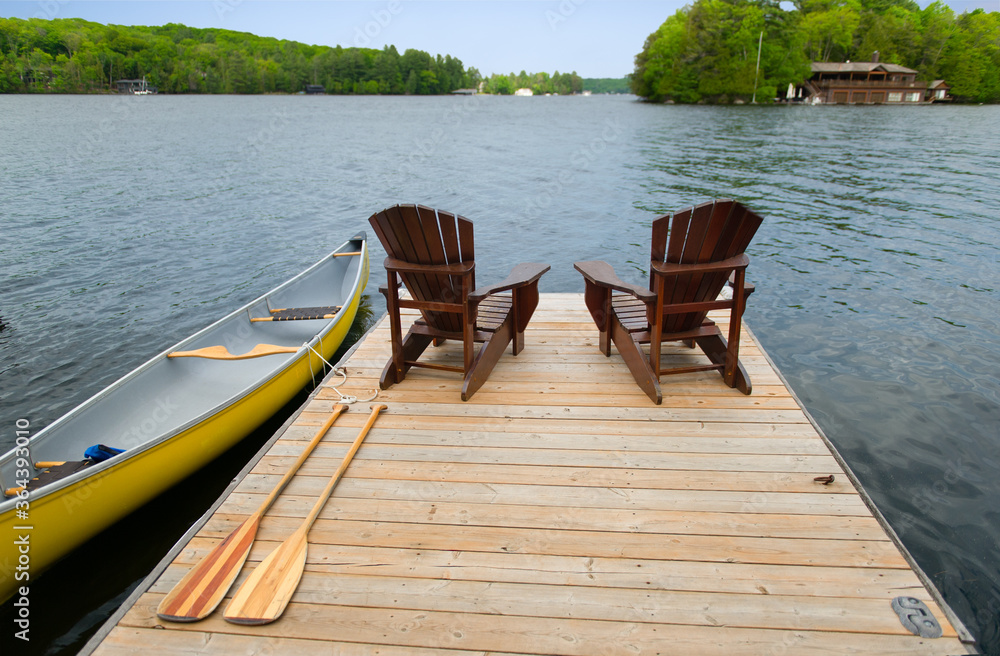 Two Adirondack chairs on a wooden dock facing the blue water of a lake in Muskoka, Ontario Canada. A yellow canoe is tied to the dock next to the oars.
