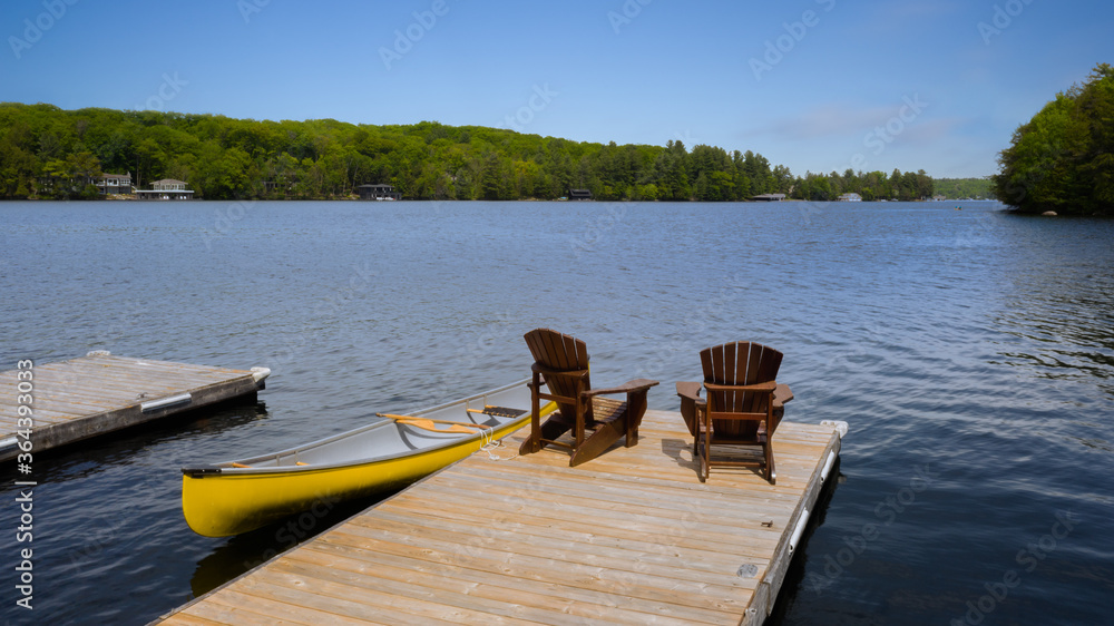 Two Adirondack chairs on a wooden dock facing a calm lake in Muskoka, Ontario Canada. A yellow canoe is tied to the dock. A cottage nestled between trees is visible across the water.