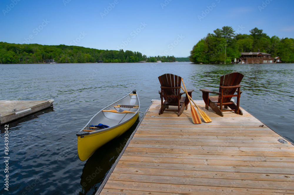 Two Adirondack chairs on a wooden dock facing the blue water of a lake in Muskoka, Ontario Canada. A yellow canoe is tied to the dock while the paddles are next to the chairs.
