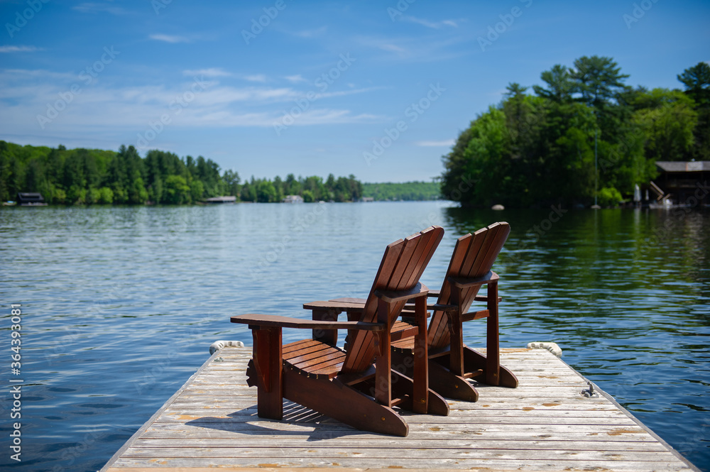 Two Adirondack chairs on a wooden dock overlooking a calm lake.  A cottage nestled between green trees is visible across the water.