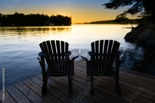 Silhouette of two Muskoka chairs sitting on a cottage wooden dock at sunset. The warm orange colours are contrasting with the blue sky.  © AC Photography