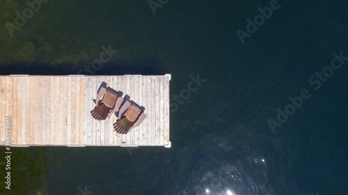 Drone aerial view of Two Adirondack chairs on a wooden dock overlooking a the calm water of a lake in Muskoka, Ontario Canada.
