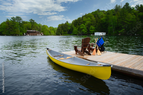 Two Adirondack chairs on a wooden dock facing the blue water of a lake in Muskoka, Ontario Canada. A yellow canoe is tied to the dock. Life jacket and oars are visible near the chairs. 