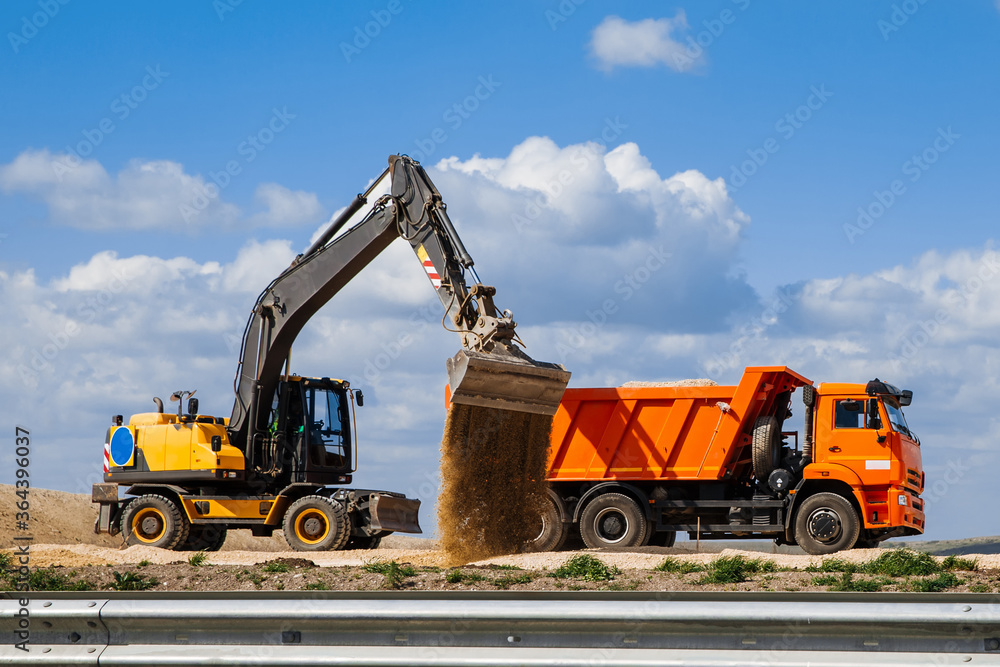 A yellow backhoe loader loads the earth into a truck during the construction of a road against a blue sky with clouds. A yellow backhoe loader loads the earth into a truck during the construction of a road against a blue sky with clouds.