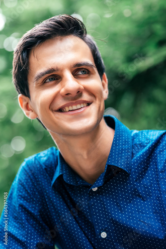 Portrait of young attractive man with blue eyes and dark hair, wearing blue shirt looking away, smiling while posing outdoors. Vertical shot © Adamov