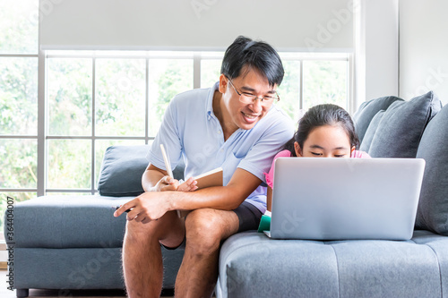 daughter learning from home with laptop on sofa in living room while father reading a book. asian family spending time together. © jirakit