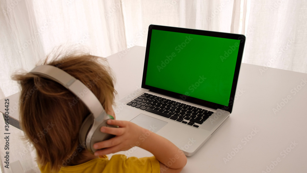 Little boy in headphones with child sits at desk looks at laptop green screen back view Little boy in headphones with child sits at desk looks at laptop green screen back view