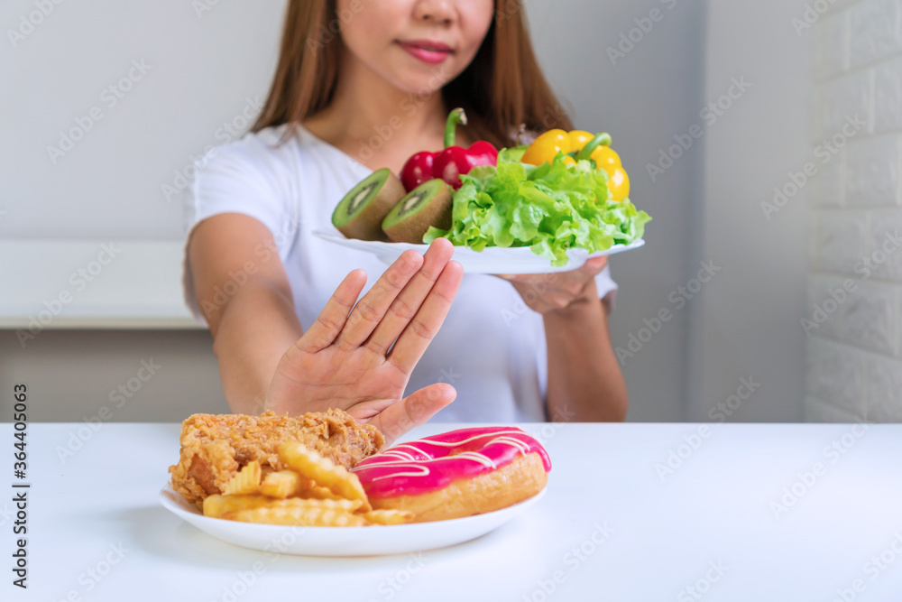 Close up of young Asian woman using hand push out her favorite fried chicken, french fries, doughnut and choose fruits, vegetables for good health. Clean eating, diet, lose weight concept. Close up