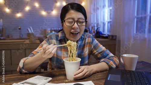 beautiful young asian woman eating instant noodles with chopsticks while working on laptop computer in night home kitchen. smiling lady with ramen soup for bedtime snack enjoy and laughing in evening