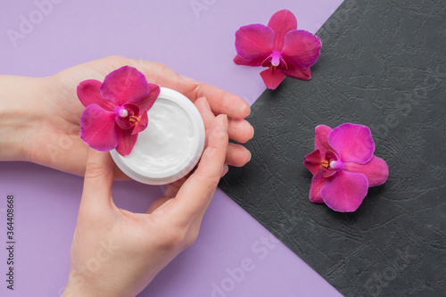 Cosmetic background with hands, soft white cream and fragile magenta colored orchids.Moisturizing facial cream in a jar and blooming orchid flowers on purple and black background, skin care cosmetics.