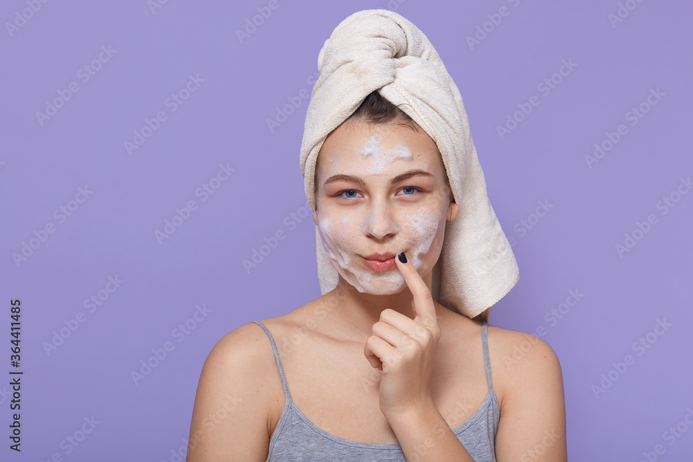 Image of lovely woman with thoughtful expression, keeps finger near corner of lips, looks at camera, applies facial mask for refreshing, wrapped in soft towel. Image of lovely woman with thoughtful expression, keeps finger near corner of lips, looks at camera, applies facial mask for refreshing, wrapped in soft towel.