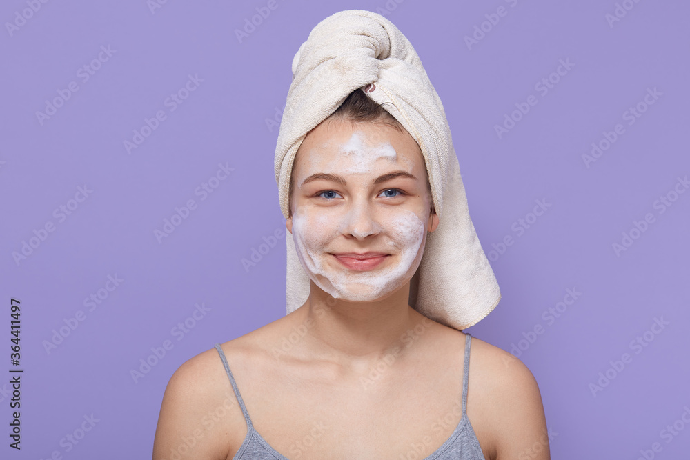 Woman with pleasant appearance, being wrapped in white towel and white facial mask on face, female posing with charming smile against lilac background. Woman with pleasant appearance, being wrapped in white towel and white facial mask on face, female posing with charming smile against lilac background.