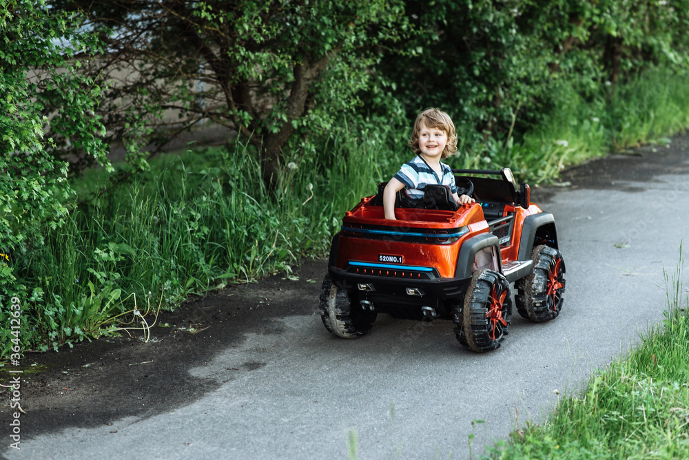 curly-haired boy in a striped T-shirt rides a red big toy car driving on an asphalt path. day off, outdoor recreation curly-haired boy in a striped T-shirt rides a red big toy car driving on an asphalt path. day off, outdoor recreation