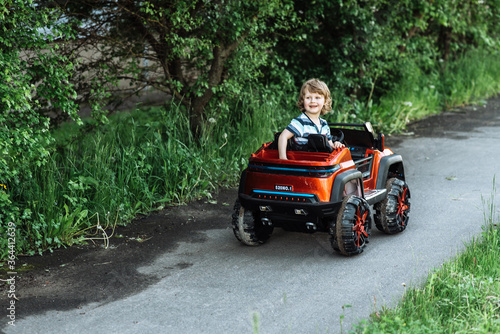 curly-haired boy in a striped T-shirt rides a red big toy car driving on an asphalt path. day off, outdoor recreation