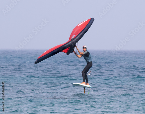 A man is wing foiling using handheld inflatable wings and hydrofoil surfboards in a blue ocean, this is a new wind sport that is becoming very popular quickly.