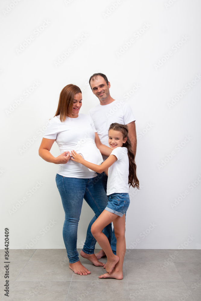 Pregnancy. Happy Family Expecting Baby. Indoor portrait of wife and husband with little laughing girl wearing casual clothers (blue jeans and white t-shirt). Pregnant mother, father and daughter Pregnancy. Happy Family Expecting Baby. Indoor portrait of wife and husband with little laughing girl wearing casual clothers (blue jeans and white t-shirt). Pregnant mother, father and daughter