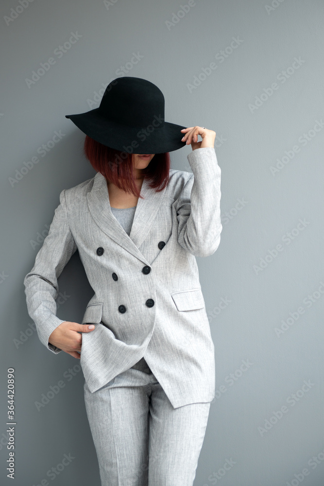 Beautiful fashion woman. Full length portrait of beautiful girl wearing light gray suit and black hat posing indoor over deep gray wall Beautiful fashion woman. Full length portrait of beautiful girl wearing light gray suit and black hat posing indoor over deep gray wall