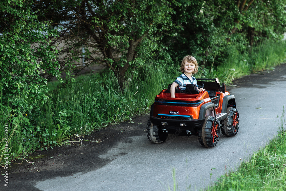 curly-haired boy in a striped T-shirt rides a red big toy car driving on an asphalt path. day off, outdoor recreation