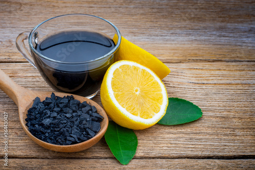Canvastavla Glass of black lemonade and broken activated charcoal powder in wooden spoon with fresh yellow lemon isolated on rustic wood table background