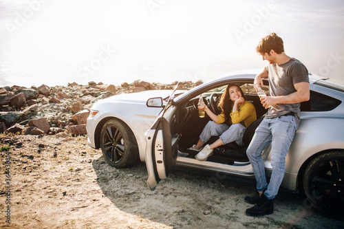 Romantic couple is standing near a Muscle car on the beach. The handsome bearded man and an attractive young woman have a Love story. © Vlad