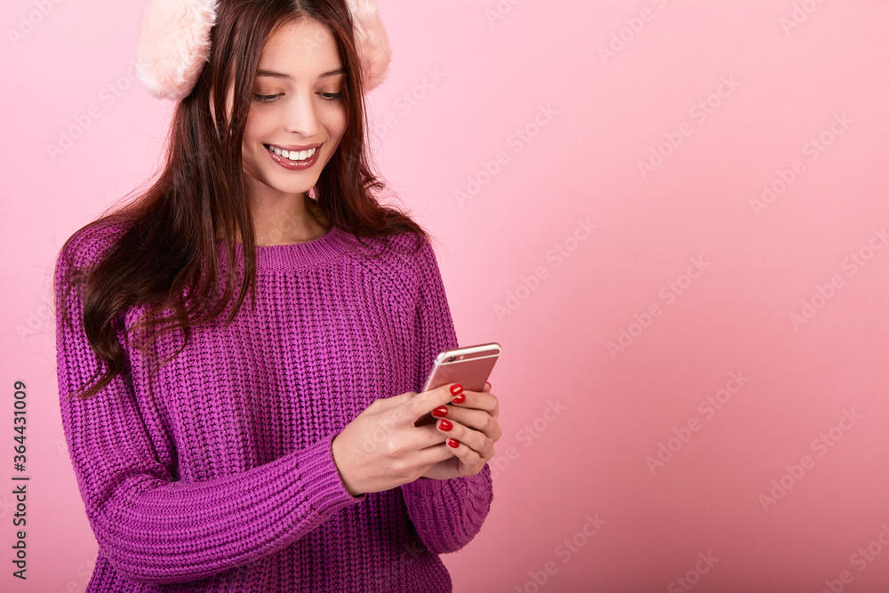 Simatic lady in a knitted sweater and winter headphones and a phone on a pink background. Simatic lady in a knitted sweater and winter headphones and a phone on a pink background.