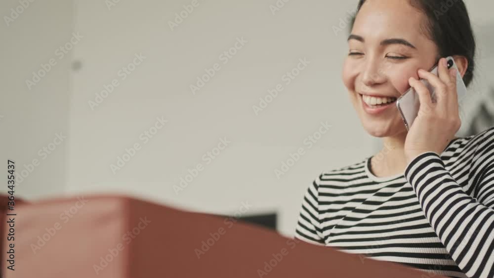 Cheerful pretty asian woman talking by smartphone and looking away while sitting on sofa at home Cheerful pretty asian woman talking by smartphone and looking away while sitting on sofa at home