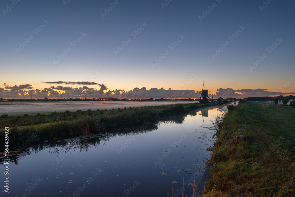 Typical dutch scene after sunset with windmill along the waterside and fog over the fields