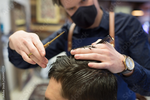 grooming, hairdressing and health safety concept - close up of male client and hairdresser wearing face medical mask for protection from virus disease with comb and scissors cutting hair at barbershop
