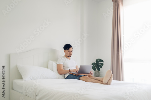 Asian man lying on bed work on his laptop in cozy white bedroom.