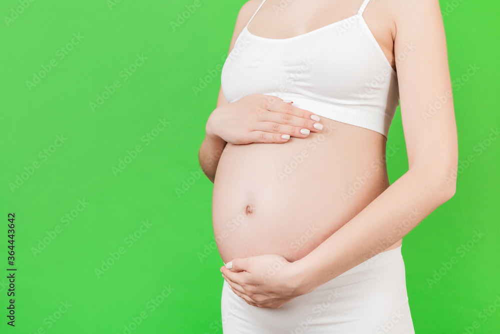 Cropped image of pregnant woman's hands embracing her abdomen at green background. Future mom is wearing white underwear. Expecting of a baby. Copy space Cropped image of pregnant woman's hands embracing her abdomen at green background. Future mom is wearing white underwear. Expecting of a baby. Copy space