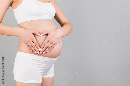 Close up of young pregnant woman making heart shape with her hands on the belly at gray background. Love to the future baby. Copy space © sosiukin